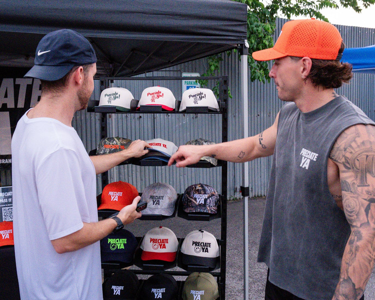 Two men standing outside under a black canopy next to a display rack of custom snapback hats. One man on the left wears a white t-shirt and blue shorts, while the other on the right wears an orange cap, a gray sleeveless shirt, and black shorts.